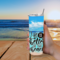 Woman holding tumbler with Life Is Better quote; her hand on table, beach scene in background, relaxing ocean vibes.
